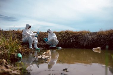 Bilim adamları ya da biyologlardan oluşan bir ekip, kimyasal dolu çöp şişeleri olan doğal bir su kaynağından su örnekleri toplamak için koruyucu giysiler giyer. Su kirliliği kavramı.