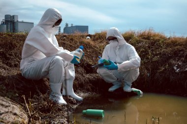 Bilim adamları ya da biyologlardan oluşan bir ekip, kimyasal dolu çöp şişeleri olan doğal bir su kaynağından su örnekleri toplamak için koruyucu giysiler giyer. Su kirliliği kavramı.