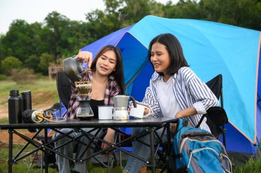 Asyalı genç iki kadın kamp çadırının yanında kahve içmeyi, kahve içmeyi ve yaz tatilinde dinlenmeyi seviyorlar..