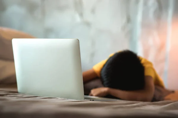 Asian kid lies down and watches social media on laptop until tired and falls asleep on the bed near electronic devices. Selective focus on laptop with Copy space. Social media digital detox Concept.