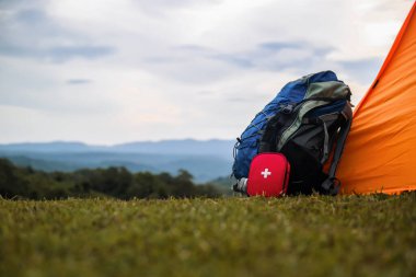 Sırt çantası ve kırmızı ilk yardım çantası çimenli bir tepede turuncu bir çadırın yanında dinleniyor, dinleniyor ve iyileşiyor dağları olan bir vücut ve arka planda bulutlu bir gökyüzü, açık hava macerasını ima ediyor..