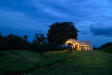 Sıcak ışıklarla aydınlatılmış rahat bir çadır akşam gökyüzünün altındaki çimenli bir tepede duruyor. Ağaçlarla çevrili sahne, doğada huzurlu ve tenha hissettiriyor..