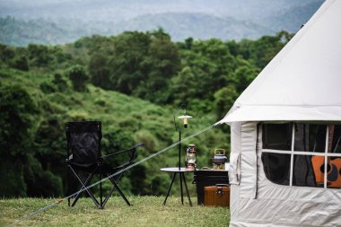 Çadırı, sandalyesi ve çimenli bir tepeye kurulmuş küçük bir masası olan huzurlu bir kamp alanı. Bir fener, çaydanlık ve diğer kamp malzemeleri arka planda yemyeşil bir orman ve dağlar ile düzenlenir..