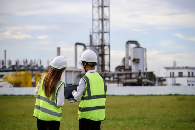 Yansıtıcı yelekli ve kasklı iki mühendis, bir sanayi fabrikasını incelerken açık hava ortamında dizüstü bilgisayarda çalışma konusunu tartışıyor. Arka planda borular ve makineler görünür..