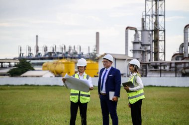 Üç mühendis sert şapka ve yansıtıcı yelek giyerek sanayi merkezinin yakınındaki boş bir arsada duruyor. Planları gözden geçiriyor ve fabrika genişlemesini tartışıyorlar..