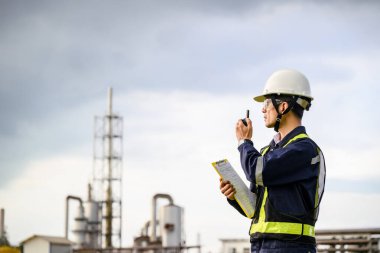 Miğfer ve yansıtıcı yelek giyen bir mühendis, bir sanayi bölgesinde pano tutarken telsiz aracılığıyla iletişim kuruyor. Arka planda makine ve kuleler görünür..