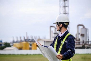 Kask takan, güvenlik gözlüğü takan ve yansıtıcı yelek giyen bir mühendis sanayi bölgesinde diz çökerken projeleri gözden geçirir. Arka planda makine ve boru tesisatı yapıları görünür