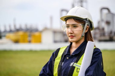 Asian female engineer, wearing a helmet, safety goggles, and a reflective vest, holds blueprints while standing in front of an industrial site. Machinery and piping structures are visible in the background. clipart