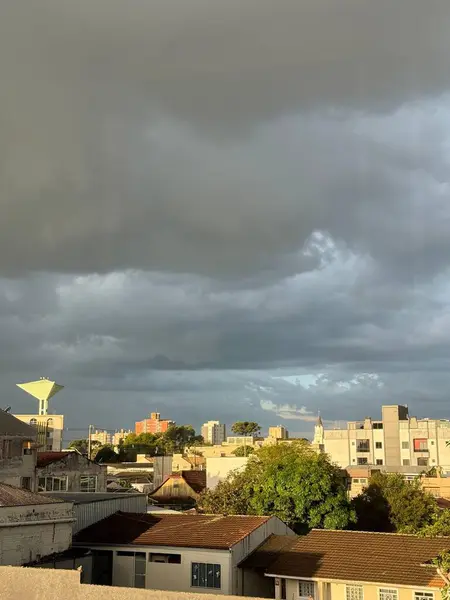 Karanlık bulutların toplandığı bulutlu bir gökyüzünün altında şehrin panoramik görüntüsü binaların ve yeşil alanların ortasındaki güneş ışığını yansıtıyor. Kentsel yapılar ve doğa arasındaki zıtlık çarpıcı, bir beklenti ve gizem ortamı yaratıyor..