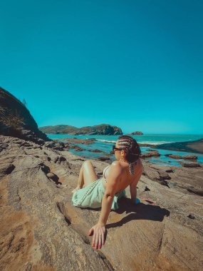 Saçlarının bir tarafında örgüleri olan bir kadın, kısa saçlı, denizin kenarında kayalarda güneşlenen güneşlenen güneşli bir günde, dağların arasında, mavi gökyüzü olan bir kumsalda.