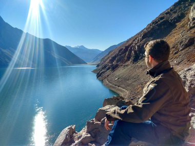 Bir kayanın üzerinde oturan, ceket ve güneş gözlüğü takan bir adam, mavi suyu olan bir barajın güzel manzarasını ve güneşli, güneşli bir günde güneşten yansıyan ışığı düşünür..