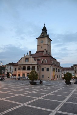 A picturesque view of the Braov Council House in Romania, with its clock tower and medieval architecture, set against an evening sky in the historic town square. clipart