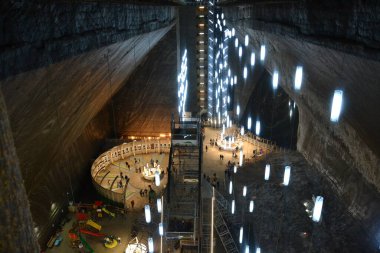 Fotoğraf Turda Tuz Madeni 'ni gösteriyor. Tavandan sarkan parlak ışıklarla dolu geniş bir yeraltı alanı. Sergide bir dönme dolap, bir oyun alanı ve eşsiz bir turizm merkezi oluşturan dairesel platformlar yer alıyor..