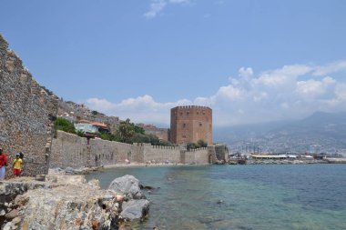 Fotoğraf, Alanya 'nın Akdeniz turkuazına bakan önemli bir ortaçağ yapısı olan Kızıl Kulesi' ni çekiyor. Taş duvarlar ve yemyeşil yeşilliklerle çevrili olan bina, tarihe ve manzaralı kıyı güzelliğine bir bakış sunuyor.