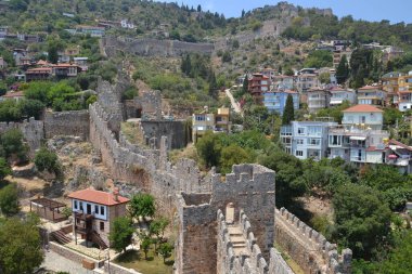 Fotoğraf Türkiye 'deki Alanya Kalesi' nin antik kalıntılarını gösteriyor. Yıkılmış taş duvarlar boyunca uzanan bir patika yeşillik, tepeler ve modern evlerle tarihi günümüz hayatıyla harmanlıyor..