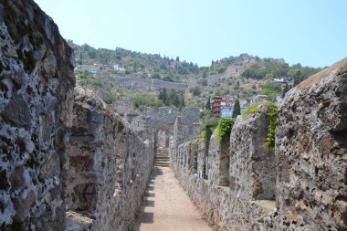 Fotoğraf Türkiye 'deki Alanya Kalesi' nin antik kalıntılarını gösteriyor. Yıkılmış taş duvarlar boyunca uzanan bir patika yeşillik, tepeler ve modern evlerle tarihi günümüz hayatıyla harmanlıyor.