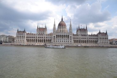 Budapeşte 'deki görkemli Macar Parlamento Binası, Neo-Gotik mimarinin çarpıcı bir örneği, Tuna Nehri kıyısında, önünde bir kayığın geçtiği parçalı bulutlu bir gökyüzünün altında gururla duruyor..