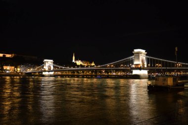 Fotoğraf Budapeşte 'nin Zincir Köprüsü' nün Tuna Nehri 'ni kaplayan gece aydınlatıldığını gösteriyor. Hilal ay cazibe katarken, su ve şehrin ışıkları çarpıcı ve ikonik bir sahne yaratıyor..