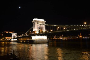 The photo shows Budapest's Chain Bridge illuminated at night, spanning the Danube River. A crescent moon adds charm, while reflections on the water and the city lights create a stunning and iconic scene. clipart