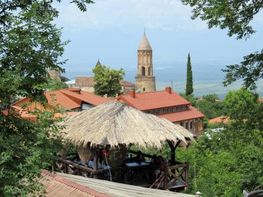 Bu fotoğraf kırmızı çatılı evleri, taştan bir kilise kulesi ve yemyeşil yemyeşil bahçeleri olan büyüleyici bir Gürcü köyünü çekiyor. Sazlı bir çardak kırsal bir dokunuş eklerken uzak vadi huzurlu bir zemin sunuyor.