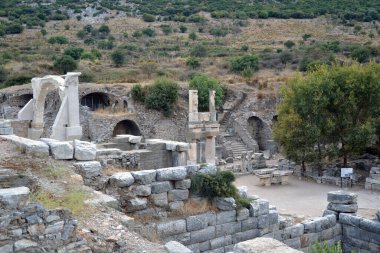 Fotoğraf, Türkiye 'nin tarihi bir kenti olan Efes' in antik kalıntılarını gösteriyor. Taş kemerler, kolonlar ve yıkılan duvarlar Roma mimarisini tasvir ediyor. Tepeler ve ağaçlarla çevrili alan zengin bir kültür mirasını yansıtıyor..