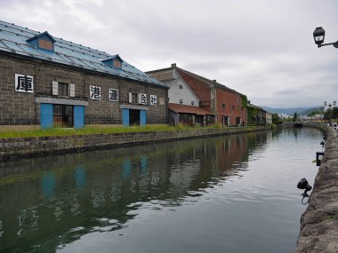 Sakin kanallar, bulutlu bir gökyüzünün altında su yüzeyine yansıyan renkli kapıları ve pencereleri olan eski tuğla depolarla ve binalarla dolu..