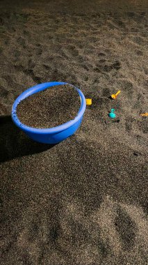 A blue plastic bucket filled with black sand, surrounded by small colorful plastic toys on a sandy surface clipart