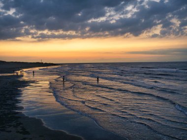 Alacakaranlıkta sakin bir sahil sahnesi. Hafif dalgalar batan güneş ışığını yansıtıyor. Dağılmış bulutlar..