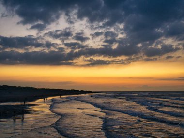 Alacakaranlıkta sakin bir sahil sahnesi. Hafif dalgalar batan güneş ışığını yansıtıyor. Dağılmış bulutlar..
