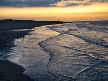 Alacakaranlıkta sakin bir sahil sahnesi. Hafif dalgalar batan güneş ışığını yansıtıyor. Dağılmış bulutlar..