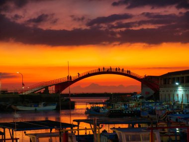 Kemerli bir köprüde insanların siluetleriyle parlayan bir günbatımı, limana yansıyan canlı gökyüzü..