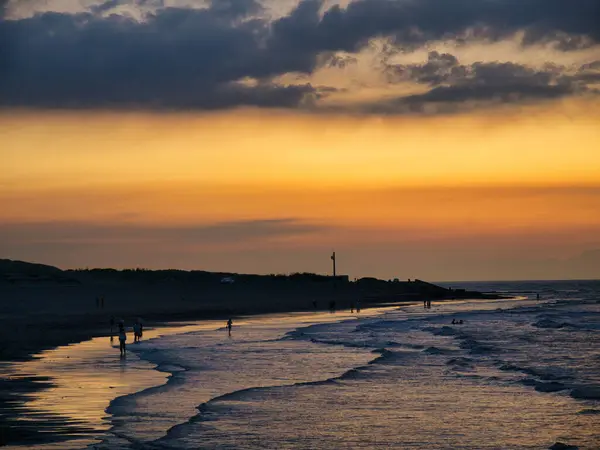 Alacakaranlıkta sakin bir sahil sahnesi. Hafif dalgalar batan güneş ışığını yansıtıyor. Dağılmış bulutlar..
