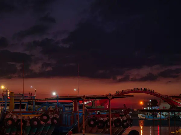 Canlı bir turuncu ve kırmızı gün batımı, sakin bir limandaki kemerli bir köprüyü ve tekneleri aydınlatarak gökyüzünü aydınlatıyor..