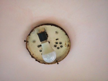 Tek bir vidalanmamış ampul paslı, dairesel tavan demirinden açık pembe bir duvara asılıyor..