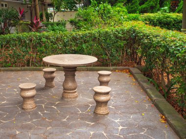 A stone table with matching stools stands on a patterned stone floor, surrounded by lush garden greenery. clipart