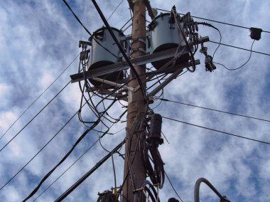 Birden fazla kablo ve trafoya sahip bir elektrik direği bulutlu bir gökyüzünün arka planına doğru uzanıyor..