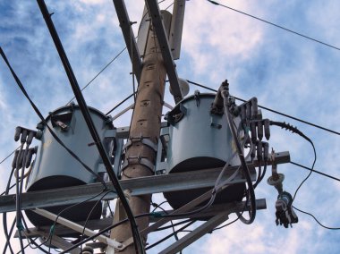 Birden fazla kablo ve trafoya sahip bir elektrik direği bulutlu bir gökyüzünün arka planına doğru uzanıyor..
