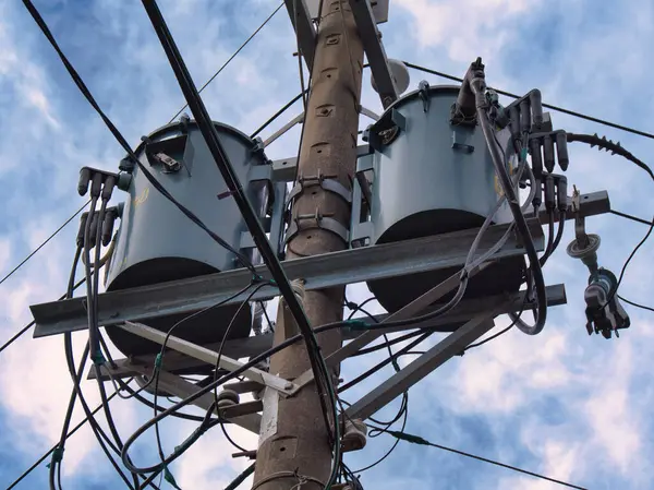 Birden fazla kablo ve trafoya sahip bir elektrik direği bulutlu bir gökyüzünün arka planına doğru uzanıyor..