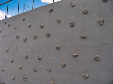 Yüzeyinde mavi bir gökyüzüne karşı dizilmiş, kayaya benzeyen çok sayıda tutacağı olan büyük, eğimli bir beton tırmanma duvarı..