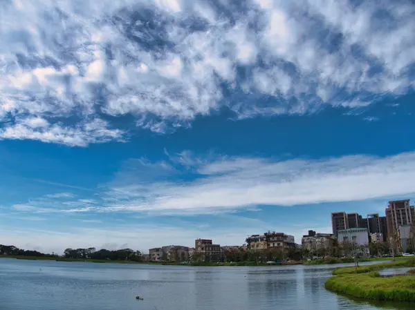 Sakin bir göl, uzak sahillerdeki binalar ve ağaçlarla çevrili, canlı, mavi bir gökyüzünün altında uzanır..