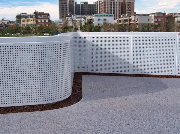 Şehir parkında peyzajlı bir parka kurulmuş kıvrımlı dizaynlı modern, beyaz delikli metal bir çit şehir binalarının arka planına karşı..