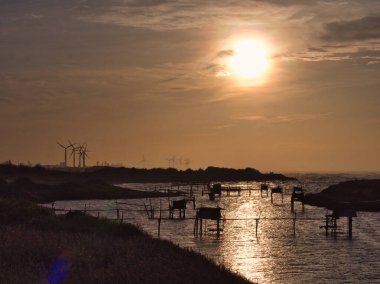 Parlak bir güneş, nehrin üzerine sıcak bir parıltı saçar. Ufukta uzak rüzgar türbinleri ve cambaz ayaklıkları üzerindeki ahşap balıkçılık yapılarını aydınlatır..