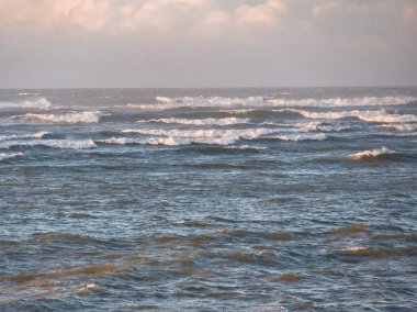 Beyaz şapkalı yumuşak dalgalar engin açık denizde hafif bulutlarla dolu yumuşak pastel bir gökyüzü ile birleşiyor..