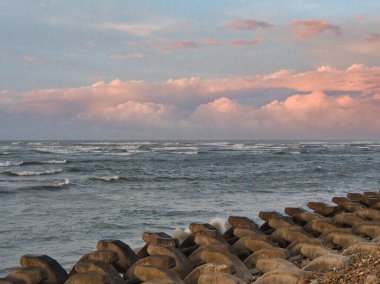 Rows of concrete wave breakers line the rocky shore, with a vast ocean stretching towards a sky filled with pink and orange clouds at dusk. clipart