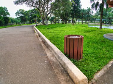 Yeşil çimlerin yanında dağınık ağaçlar ve kahverengi ahşap çöp tenekesi olan yol huzurlu ve ferah bir park ortamı yaratıyor..