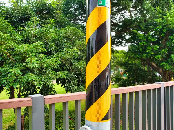Sarı ve siyah çizgili uyarı direği. Yeşillik, ağaçlar ve arka planda çimenlik olan bir parktaki korkulukların yanında..
