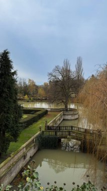 Üzerinde köprü ve arka planda ağaçlar olan bir gölet. Su bulanık ve gökyüzü bulutlu.