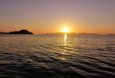 Göz kamaştırıcı altın bir günbatımı uzak dağların arkasına iniyor, sıcak renkleri gökyüzüne saçıyor ve denizin yüzeyine güzel bir şekilde yansıtıyor..