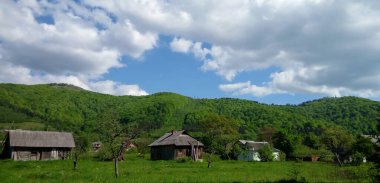 Karpat Dağları 'nın sakin kırsal kesiminde, berrak mavi gökyüzünün altında yuvarlanan yeşil tepelerin arasına yerleştirilmiş geleneksel ahşap evler yer alıyor..