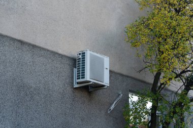 Gri bir bina duvarına monte edilmiş eski bir klima, aşınma ve yaşlanma belirtileri gösteriyor. Yıpranmış dış yüzey, duvarın düz yüzeyine karşı kontrasttır..